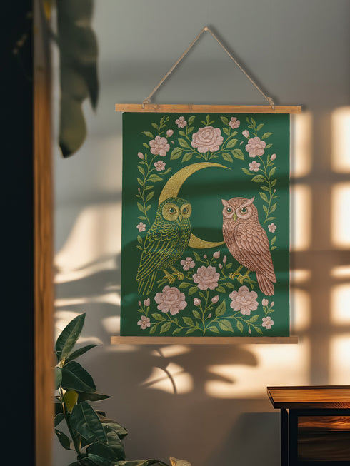 26x36 inch vertical wall tapestry printed on silky polyester, featuring a pair of embroidered-style owls perched on a golden crescent moon, surrounded by pink vintage-style florals and greenery on a forest green background. Includes stained wood slats and twine for easy hanging. A romantic, nature-inspired design for woodland and cottagecore decor lovers.